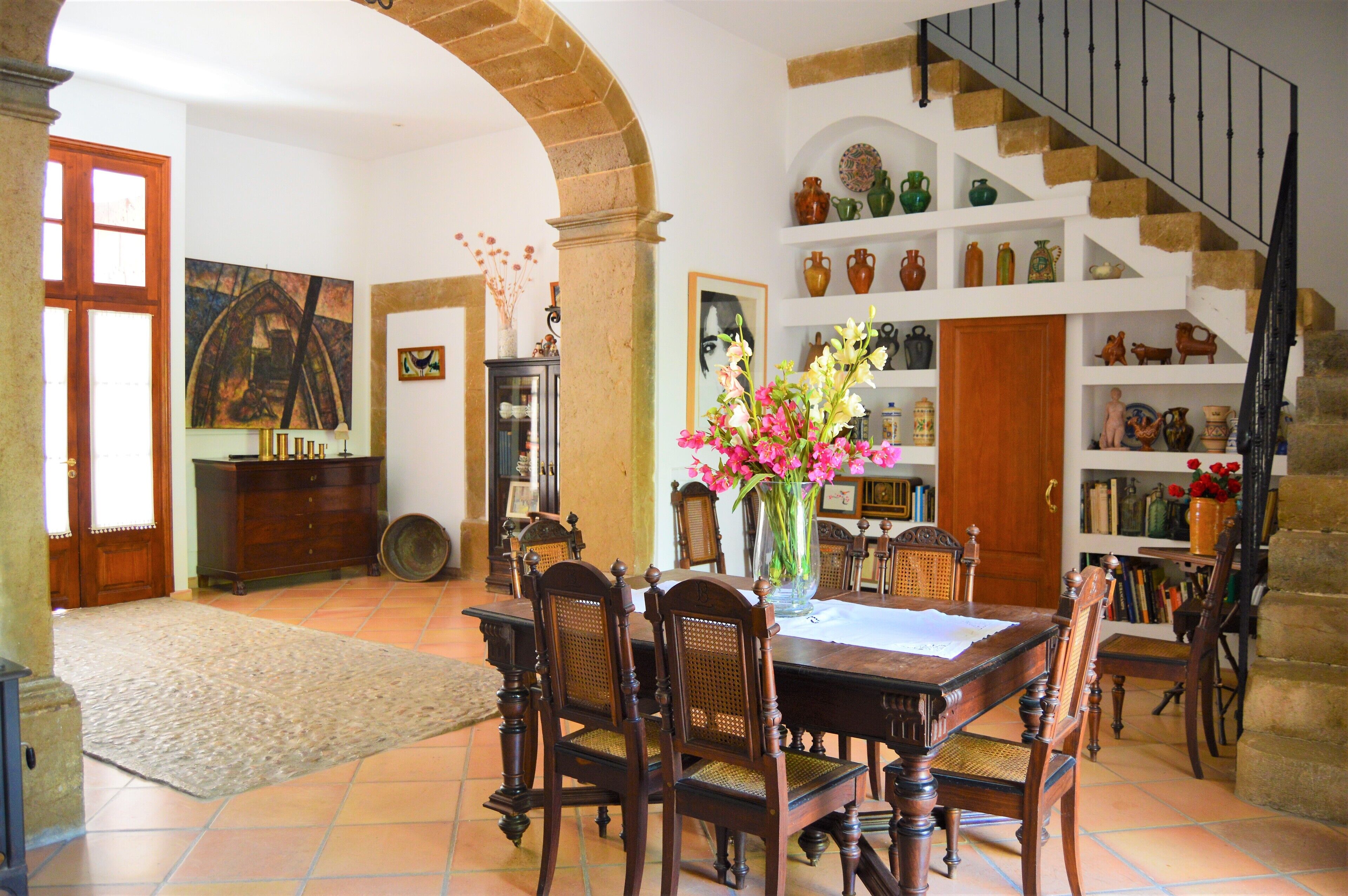 Un coin repas avec une table et des chaises en bois, un vase rempli de fleurs et un escalier orné d’étagères où sont exposées des pièces de poterie.