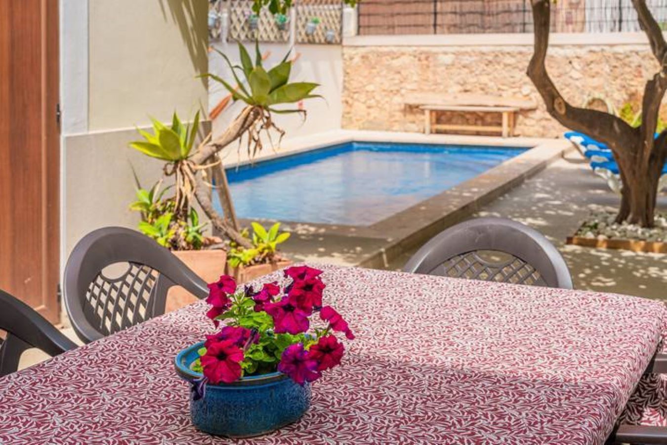 A poolside table with a patterned tablecloth, a blue ceramic pot with red flowers, and a pool in the background.