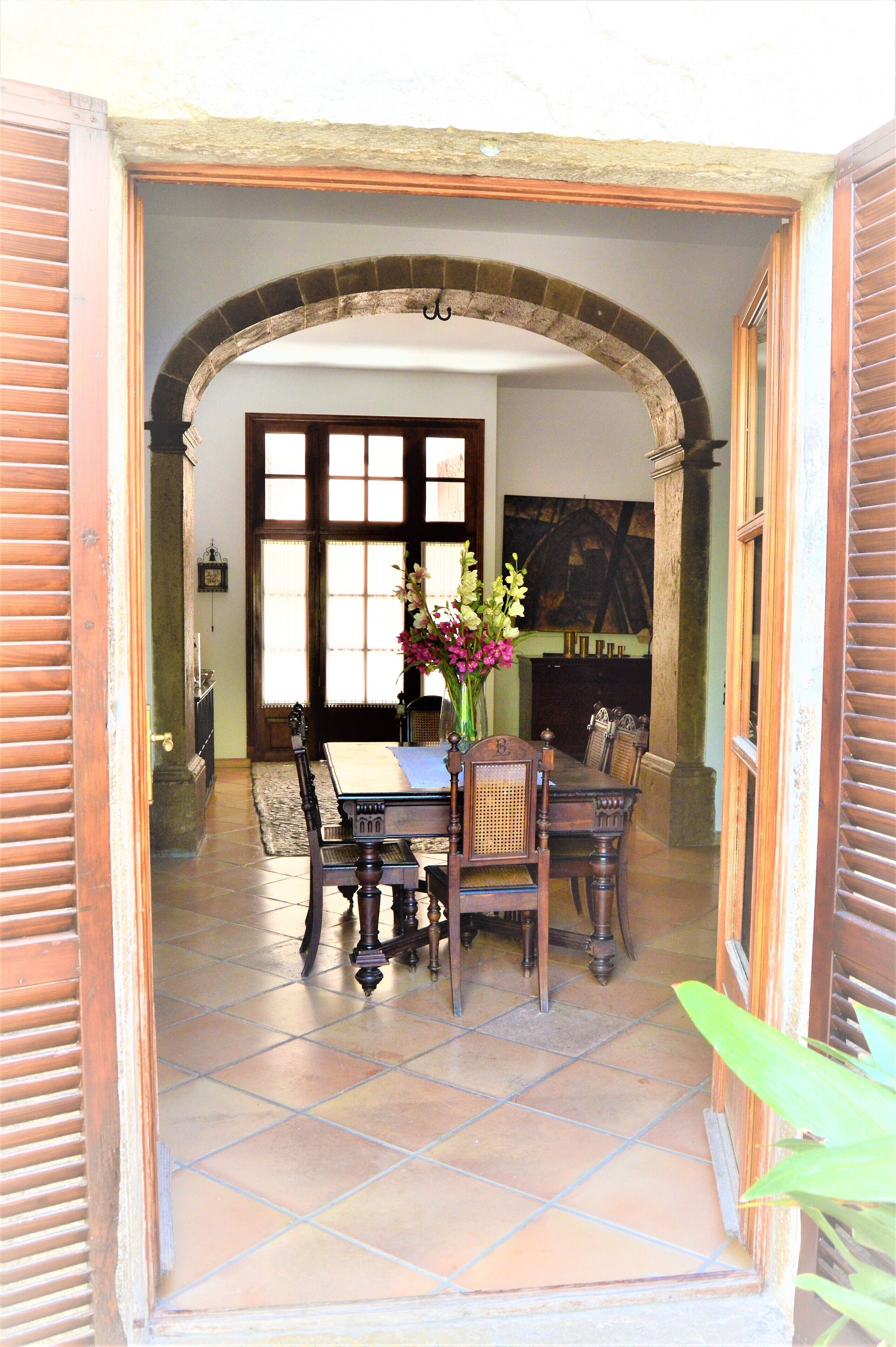 Une porte ouverte donne sur un coin repas, où l’on aperçoit une table et des chaises, un vase de fleurs et, au fond, une cheminée.