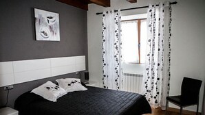 Un dormitorio con cama, una silla, mesita de noche, un cuadro en la pared y ventana con cortinas.