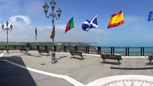Eine Uferpromenade mit Bänken und Flaggen, wolkenloser Himmel und Meerblick.