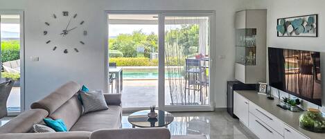 A modern living room with a sofa, a coffee table, a TV, and a sliding glass door leading to a patio with a pool.