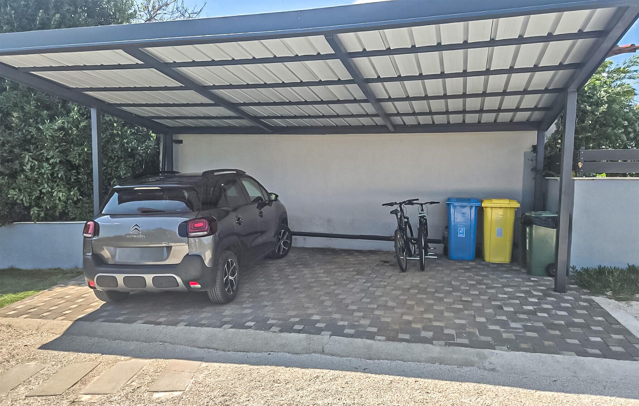 A covered carport with a parked SUV, a bicycle, and two trash bins.