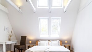 A bedroom with a large bed, two windows, a chair, a table with a book, and a bedside lamp. - Ferienhaus JohannisNest: Sauna & Kamin, große Wiese, Südterrasse mit Gasgrill (Poppenbüll)