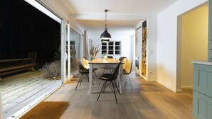 Un espace de repas moderne avec une table et des chaises en bois, un luminaire suspendu et une vue sur l’extérieur.