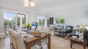 A spacious living area with a dining table set for a meal, a black leather sofa, and a TV in the background.