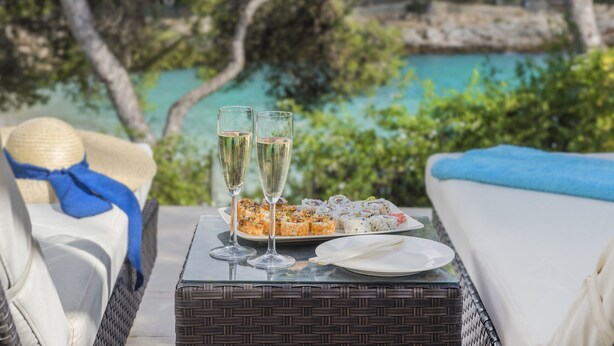 A wicker table with a tray of sushi and two glasses of champagne, set on a balcony overlooking a turquoise sea and greenery.