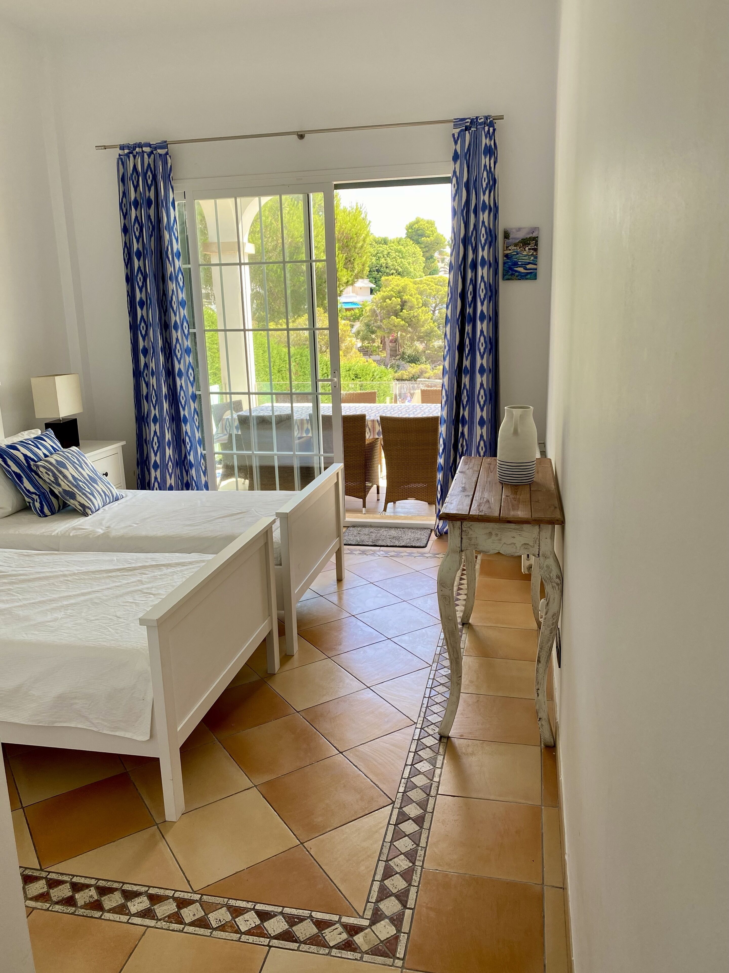 A bedroom with a bed, a nightstand, a lamp, a window with curtains, and a view of the outdoors.