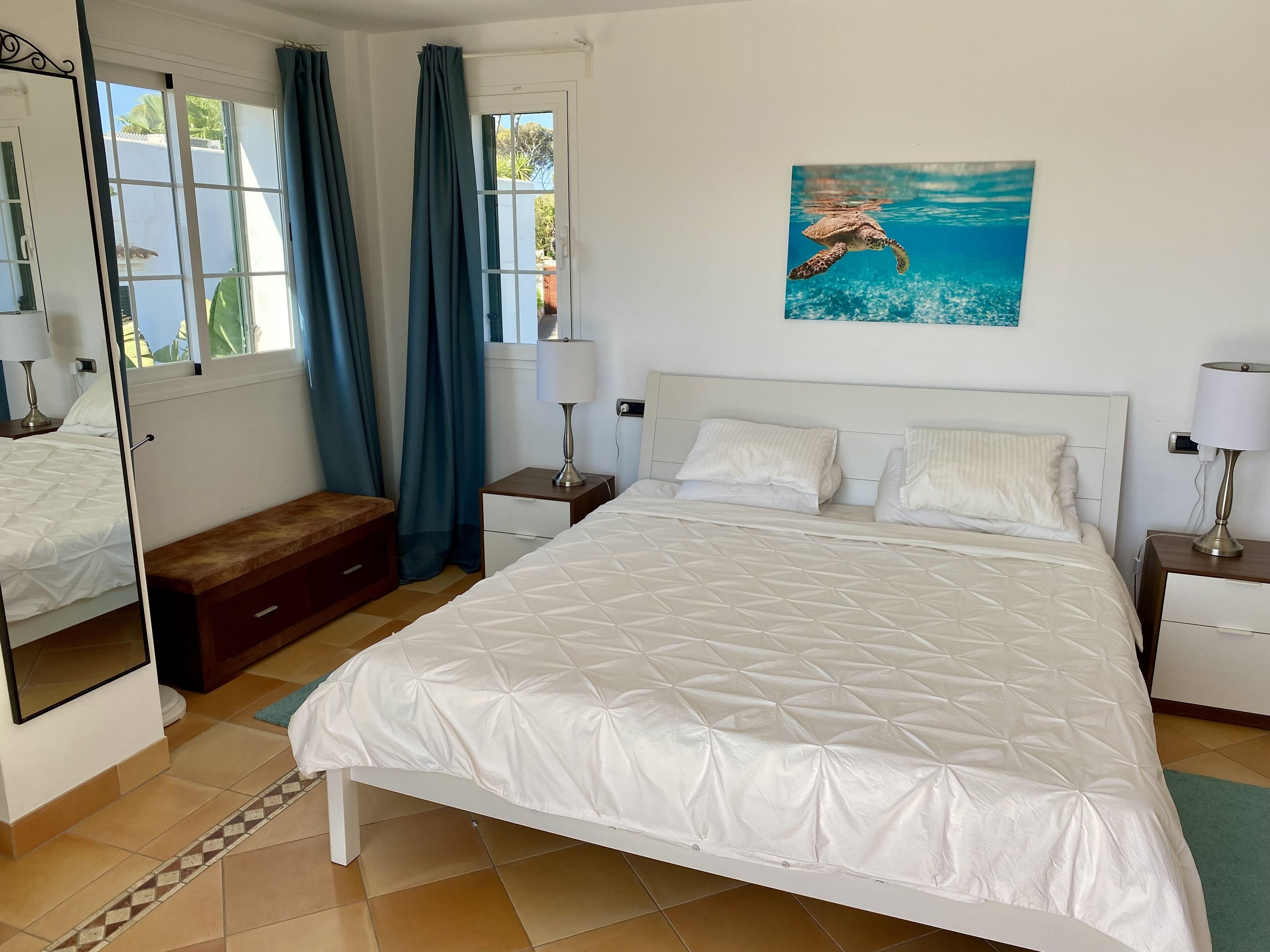 A bedroom with a large bed, a bedside table, a nightstand, a bench, a mirror, and a window with curtains.
