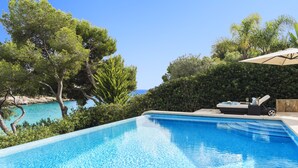 A clear blue swimming pool with a view of the sea, surrounded by lush greenery and a sun lounger.