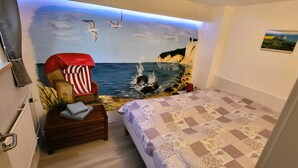 A bedroom with a mural of a beach scene, a red chair, a small table with a towel, and a bed with a patterned bedspread. - Great basement apartment in the middle of the island of Rügen in the outskirts of a village near mountains (Parchtitz)