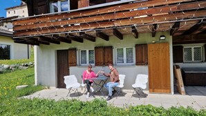 Un patio con dos sillas blancas y una mesa, una pérgola de madera en lo alto y una pared de piedra con flores amarillas.