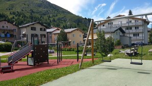 Un parque infantil con toboganes y columpios, rodeado de casas y una montaña al fondo.