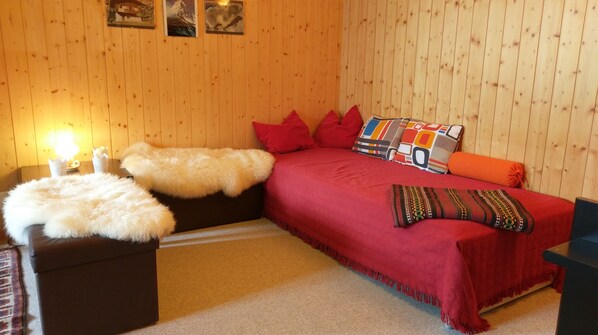 Una habitación acogedora con pared de madera, ropa de cama roja sobre la cama, un felpudo blanco y una mesita con una lámpara.