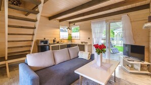 A cozy cabin interior with a sofa, a table with a vase of flowers, a television, and a kitchen area with a window. - Woodhouse Resort Dobrogoszcz (Dobrogoszcz)