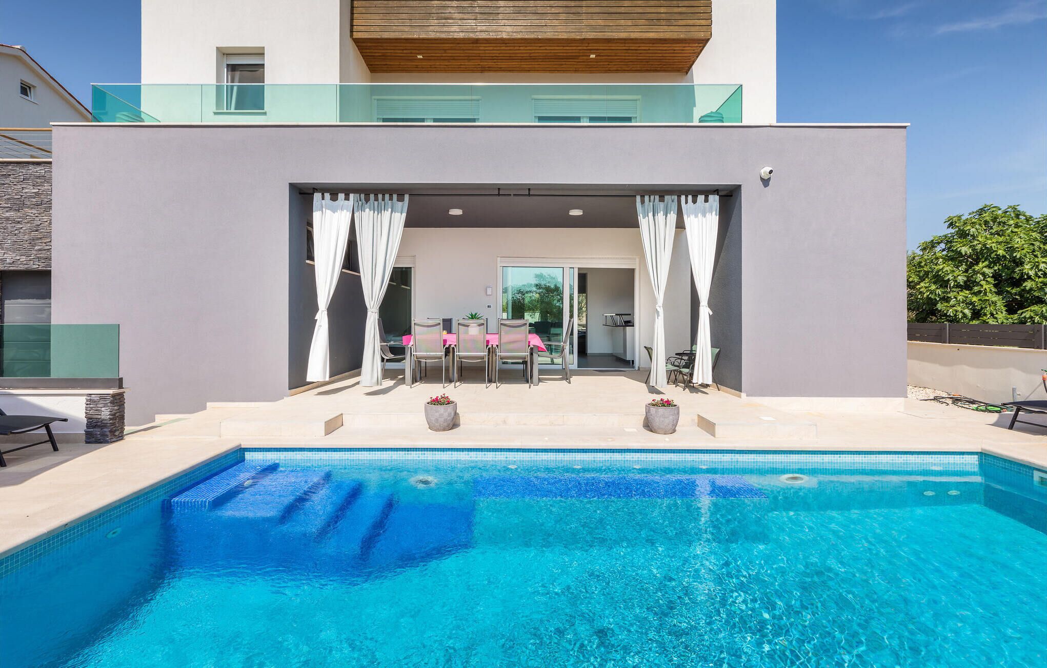 A modern pool area with a clear blue pool, a covered seating area with a table and chairs, and a view of a building with large windows and a balcony.