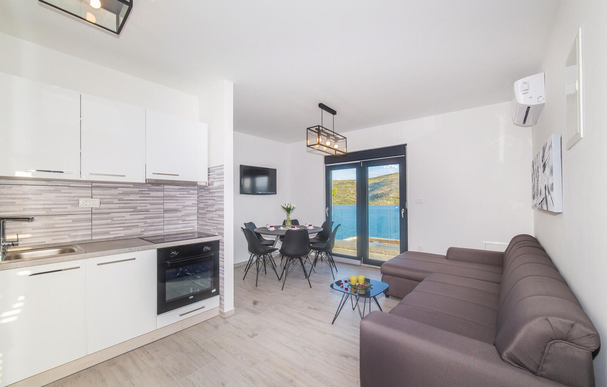 Una cocina moderna con armarios blancos, horno empotrado y salpicadero de azulejos. Hay un comedor con mesa y sillas, y un amplio ventanal con vistas al mar.