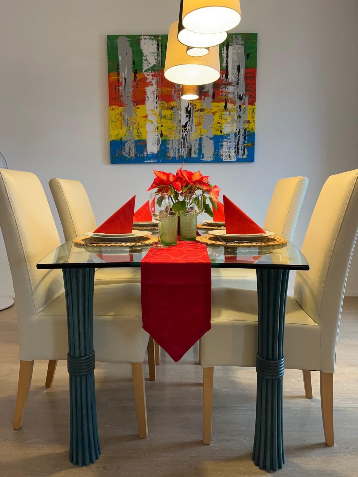Ein Esstisch mit roter Tischdecke, zwei Glasvasen mit roten Blumen und ein farbenfrohes abstraktes Gemälde an der Wand.