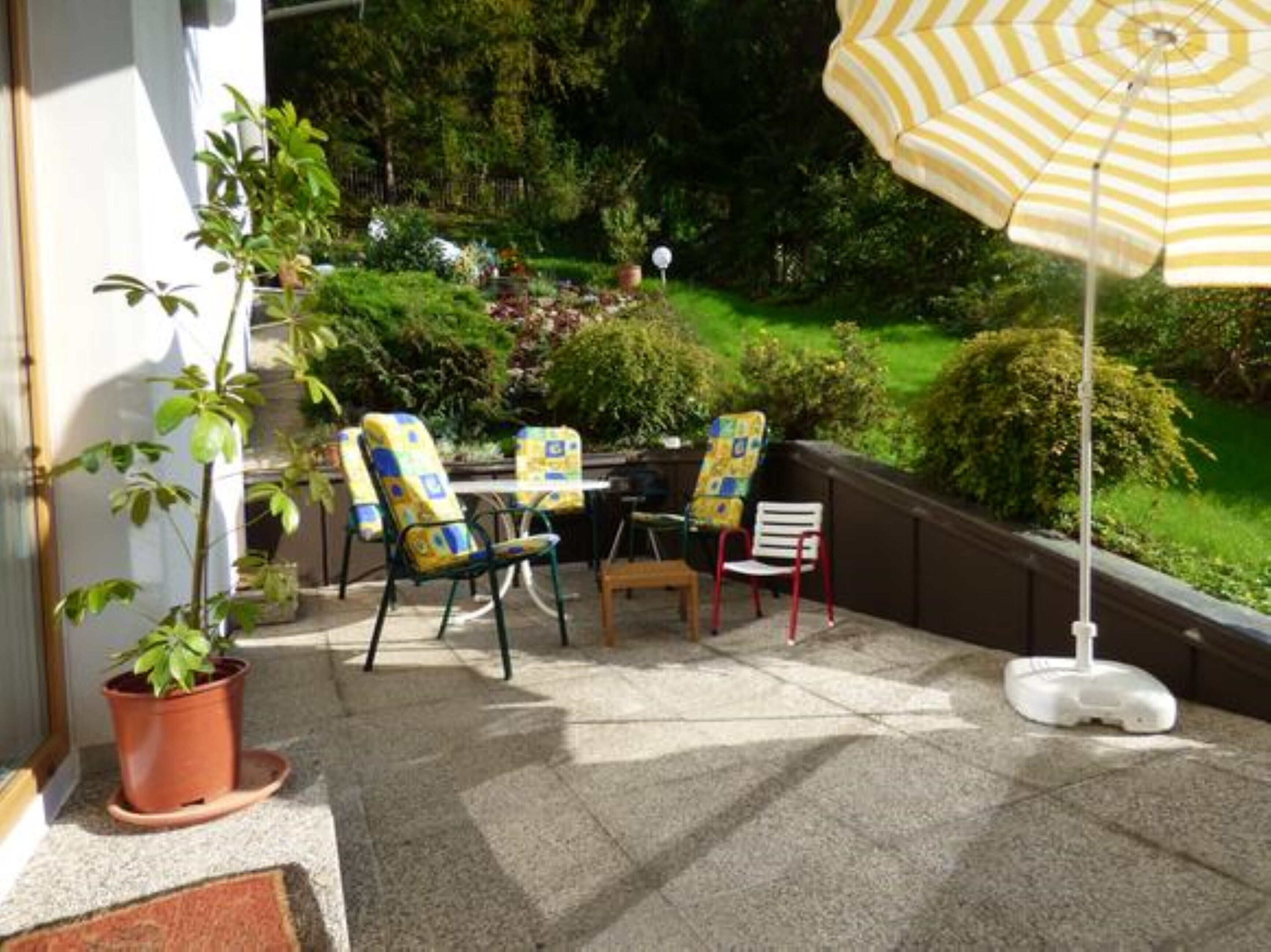 Een terras buiten met twee stoelen, een kleine tafel, een bloempot en een gestreepte parasol.