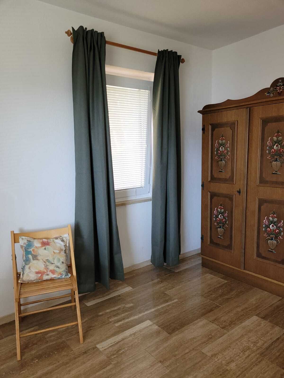 A room with a wooden wardrobe, a window with blinds, and a wooden chair with a cushion.