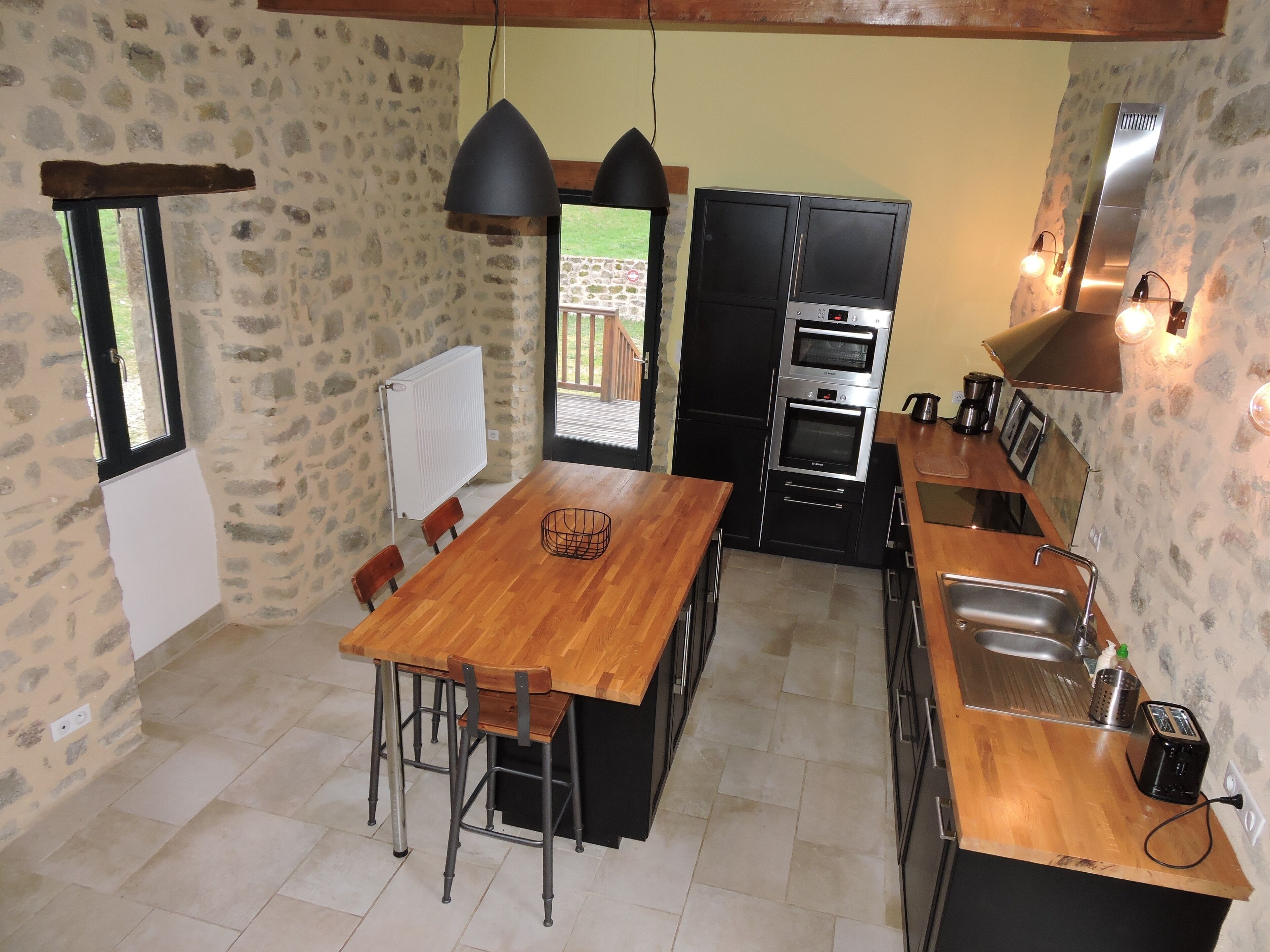 A kitchen with a wooden table, chairs, a black oven, and a stone wall.
