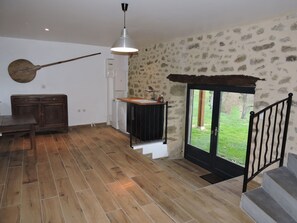 A spacious living area with a wooden floor, a dining table, a hanging light fixture, and a view through a glass door to a garden.