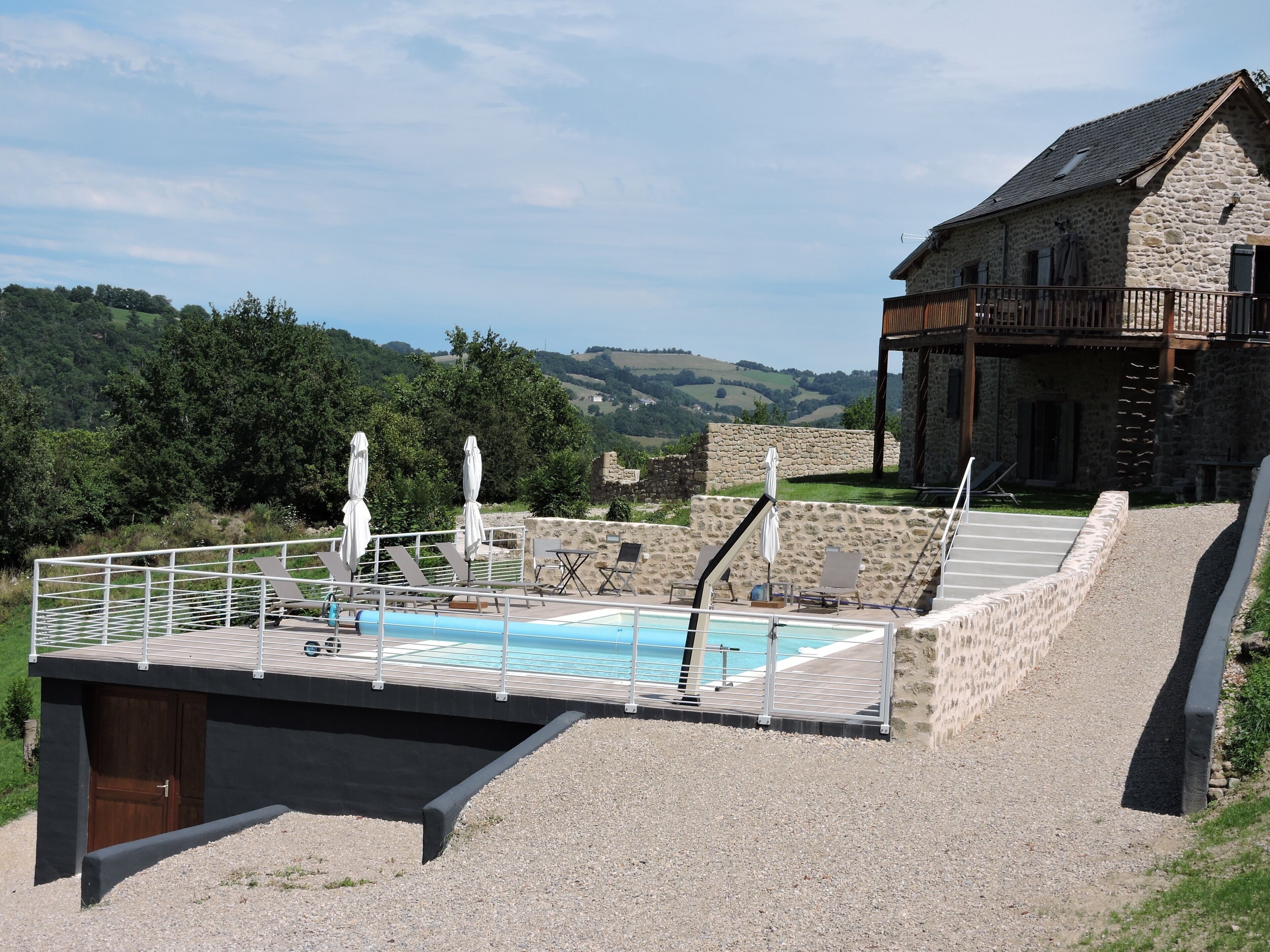 Ein Poolbereich mit Liegestühlen und Sonnenschirmen, ein Gebäude mit Steinbau und einem Balkon, sowie ein Blick auf grüne Hügel im Hintergrund.