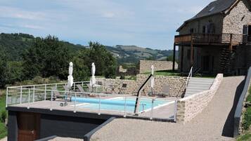 Ein Poolbereich mit Liegestühlen und Sonnenschirmen, ein Gebäude mit Steinbau und einem Balkon, sowie ein Blick auf grüne Hügel im Hintergrund.
