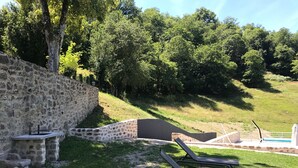 Un muro de piedra, una mesita pequeña y dos sillones de exterior en un espacio ajardinado, con árboles al fondo.