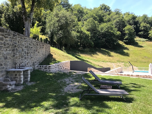 Eine steinerne Mauer, ein kleiner Tisch und zwei Liegestühle in einer grünen Außenanlage mit Bäumen im Hintergrund.