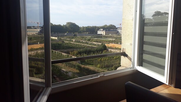 Ein Zimmer mit großem Fenster und Blick auf eine angelegte Grünfläche, im Hintergrund sind Gebäude zu sehen.