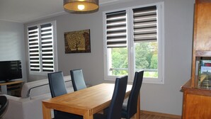 Ein Essbereich mit Holztisch und Stühlen, einer Couch, einem Fernseher und Fenstern mit Jalousien.