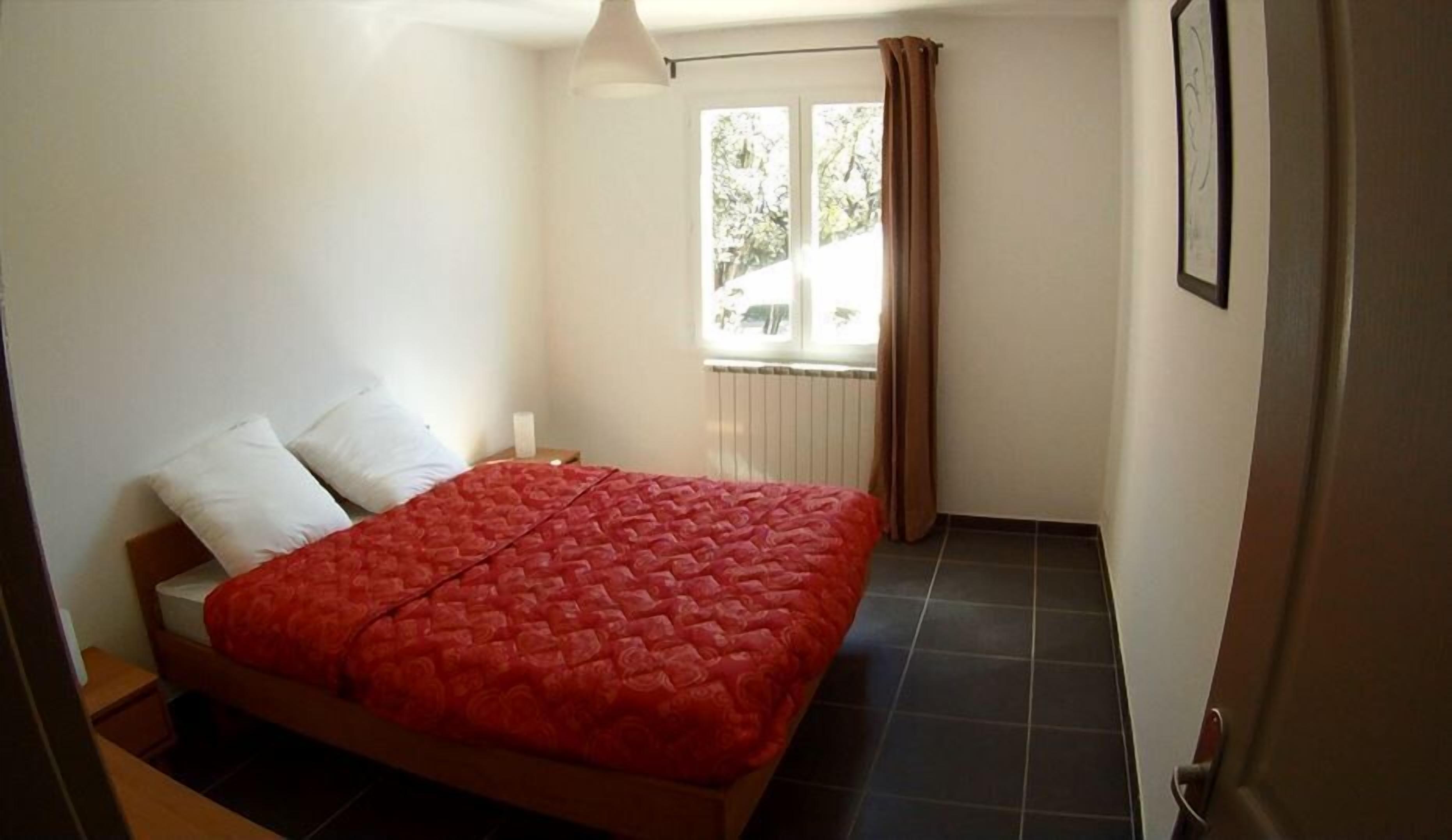 Et soveværelse med en seng, hvide puder, et rødt quiltet sengetæppe, et natbord, et vindue med gardiner og en radiator.