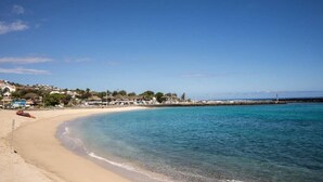 Une plage avec une eau d’un bleu limpide, un rivage sablonneux et des bâtiments en arrière-plan.