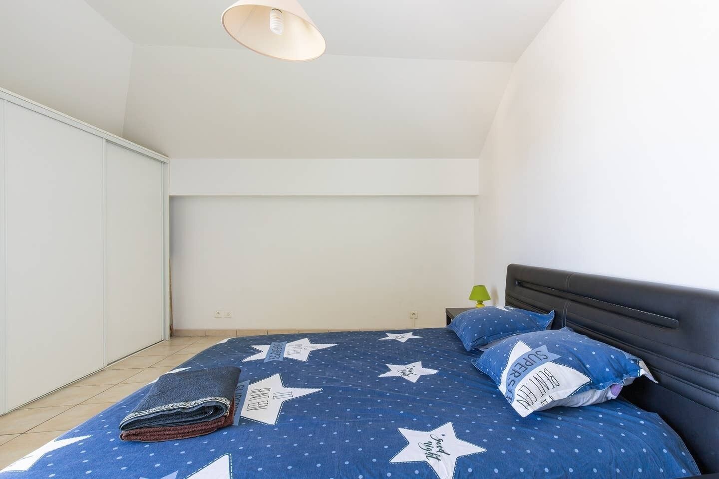 Een slaapkamer met een bed, kussens, een nachtkastje en een plafondlamp.