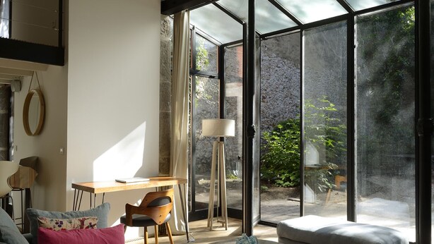 A modern living room with a glass door leading to a garden, a wooden table, and a sofa with cushions.