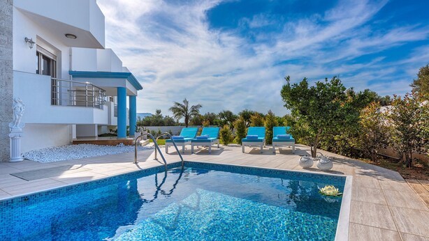 Un espace piscine moderne avec une piscine à l’eau d’un bleu limpide, un bâtiment blanc agrémenté de touches de bleu et quelques chaises longues.