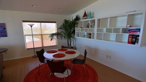 A dining area with a round table, four chairs, and a red rug. There is a window with blinds, a potted plant, and shelves with various items. - Beachfront Condo With Panoramic Views of the Gulf Plus Beach Gear, Grill & Wifi (Indian Rocks Beach)