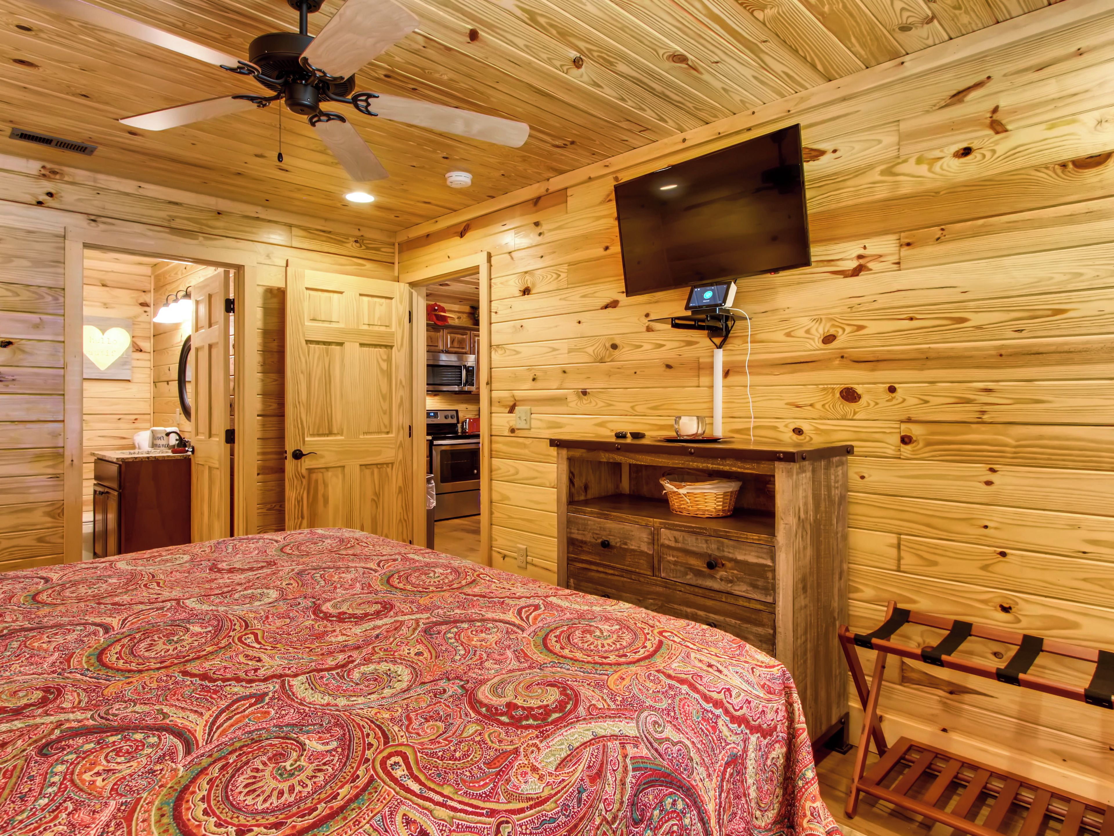 A wooden cabin room with a bed, a ceiling fan, a TV, and a small kitchenette.