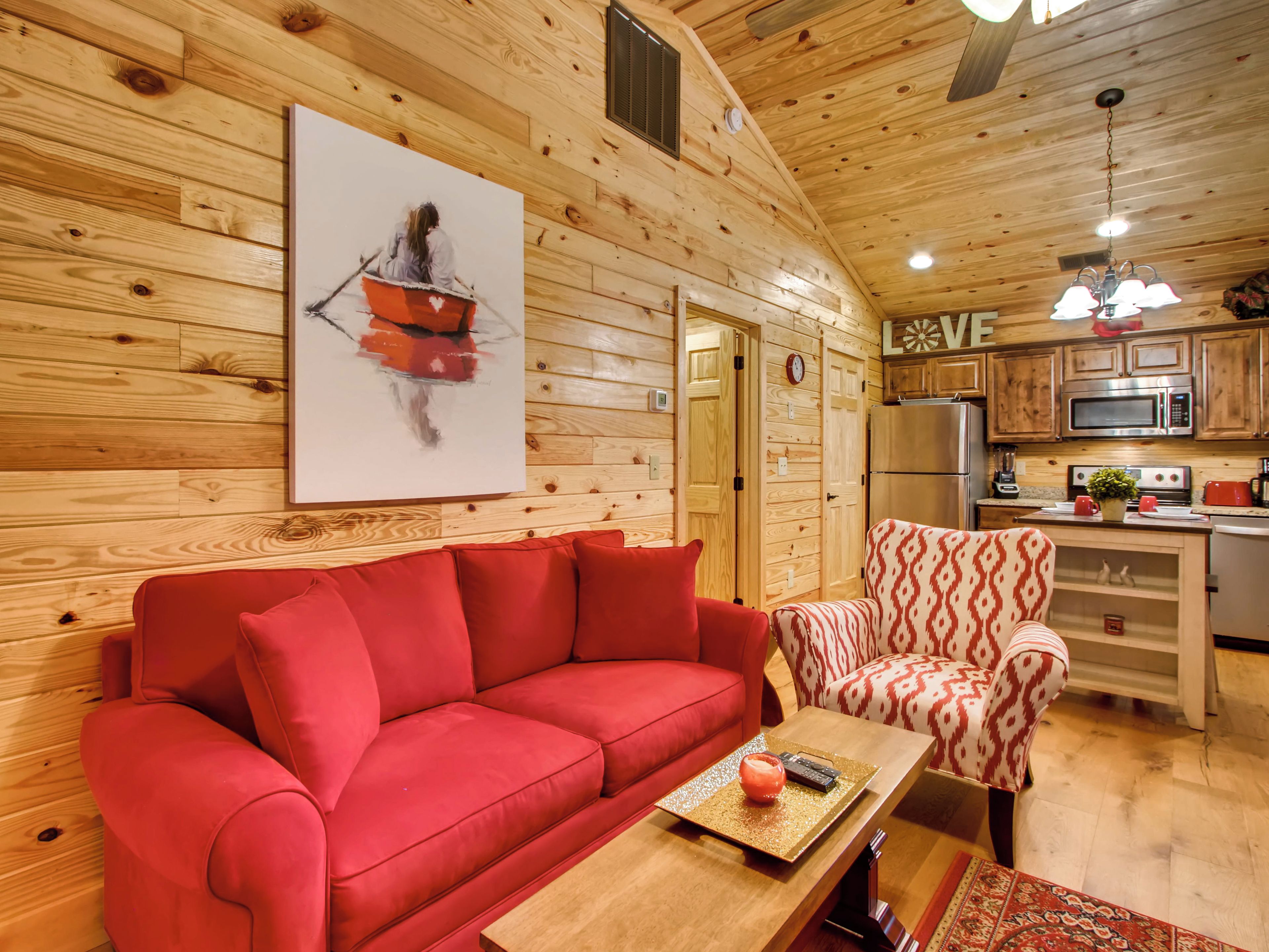 Un interior de cabaña acogedor, con un sofá rojo, un sillón estampado, paredes de madera y una zona de cocina.