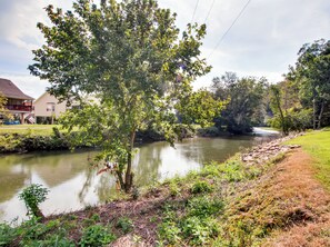 A river with a tree in the foreground, a grassy bank, and a building in the background.