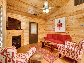 A cozy wooden cabin interior with a fireplace, a red sofa, patterned armchairs, and a ceiling fan.