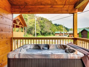 A wooden cabin with a hot tub on a wooden deck, surrounded by trees and a view of a small building in the distance.