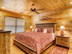 A wooden-paneled bedroom with a bed, bedside lamps, a ceiling fan, and a window with blinds. - Riverside | Pigeon Forge | Pet Friendly | Hot Tub | Arcade | WiFi (PIGEON FORGE)