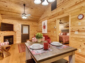 A wooden cabin interior with a dining area, a fireplace, a television, and a bed with patterned bedding.