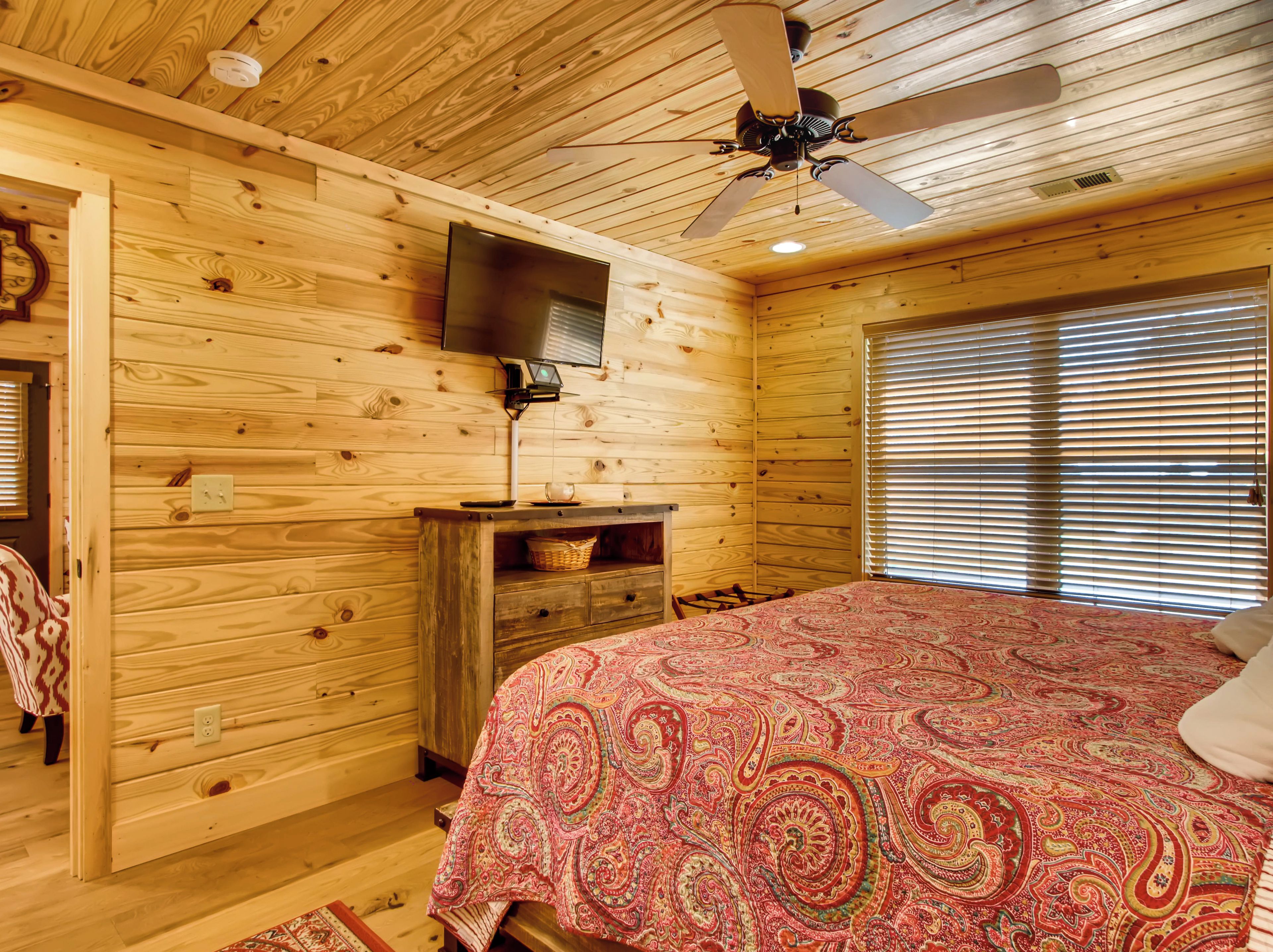 Un dormitorio de cabaña de madera con una cama, un ventilador de techo, un televisor y una ventana con persianas.