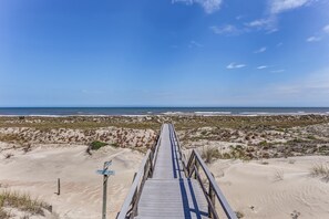 Una pasarela de madera que conduce a una playa con dunas de arena y vegetación escasa.