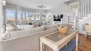 A spacious living room with a large sectional sofa, a glass coffee table, a ceiling fan, and a staircase with white railings. - Welcome to BIS BALD!, your Oceanfront Escape on BHI! (Bald Head Island)