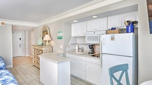 A compact kitchen with white cabinets, a microwave, and a refrigerator. There is a small island with a sink and a chair. - Oceanfront Retreat- Poolside View- Budget Friendly (Daytona Beach)