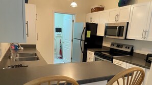 A kitchen with white cabinets, a stainless steel sink, a microwave, and a stove. There are two wooden chairs and a small island with a countertop.